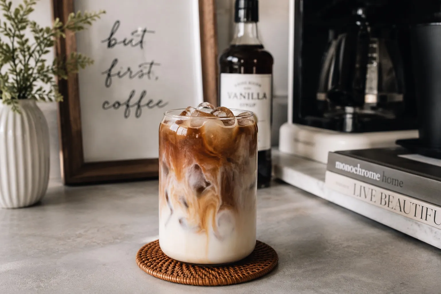 An iced coffee with a swirl of milk in a clear glass, set against a modern kitchen coffee station background.