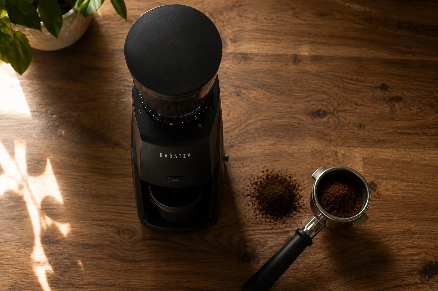 Top-down shot of a black conical burr grinder on a wooden counter in soft morning light, a small pile of fresh grounds beside the portafilter