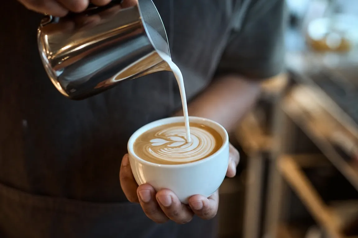 A stainless steel milk pitcher tilted mid-pour over an espresso cup, glossy microfoam catching morning light